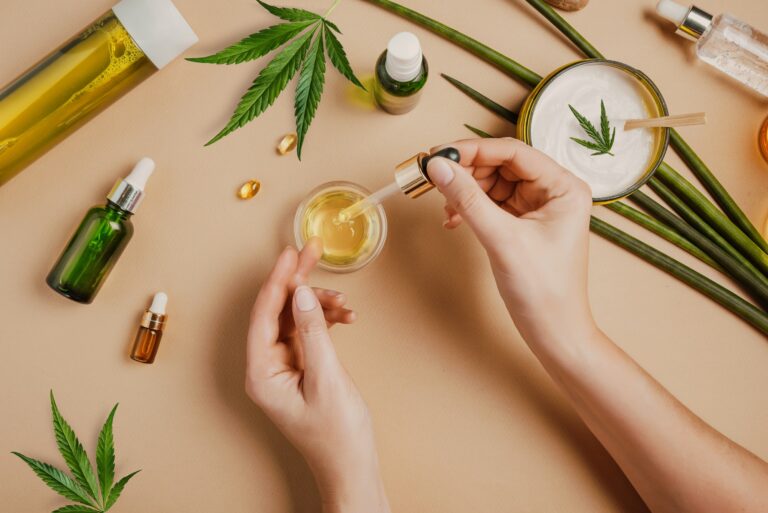 Pipette CBD cosmetic oil in female hands on background with cosmetics, cream cannabis hemp leaves